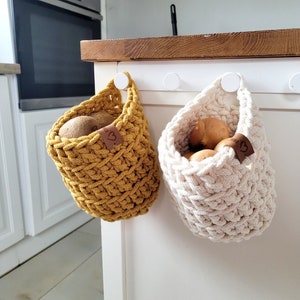 May include: Two crocheted hanging baskets with brown leather labels. The basket on the left is mustard yellow and holds kiwi fruit. The basket on the right is white and holds onions.