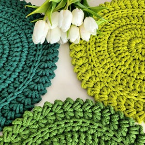 Könnte beinhalten: Nahaufnahme von drei gehäkelten Tischsets in Grüntönen mit einem Strauß weißer Tulpen. Die Tischsets sind kreisförmig und haben ein strukturiertes, geschlungenes Design. Die Farben reichen von tiefem Teal bis zu hellem Limettengrün.