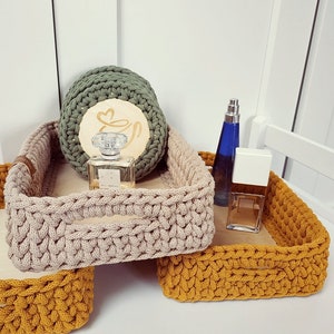 May include: Three crocheted baskets in beige, mustard yellow, and green. The baskets are stacked on top of each other. The top basket has a wooden coaster with a heart design and a bottle of perfume inside. The middle basket has a bottle of perfume and a white box. The bottom basket is empty.