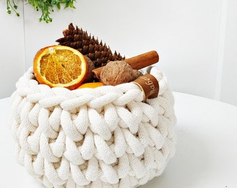 May include: A white crocheted bowl filled with oranges, cinnamon sticks, and walnuts. The bowl has a brown leather tag with the word "Baggers" on it.