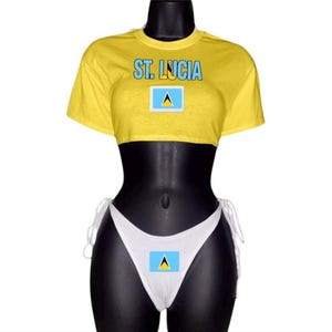May include: Yellow crop top and white bikini bottom set. The top features the text "ST. LUCIA" above a St. Lucia flag graphic. The bikini bottom also has a St. Lucia flag graphic. The set is displayed on a black mannequin.