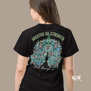 May include: Black t-shirt with a botanical lung design. The design features green leaves, pink flowers, and blue butterflies, with the text "BREATHS OF STRENGTH" and "travel through me" in green. The shirt is a crew neck.