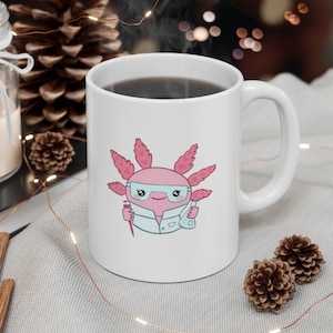 K&ouml;nnte beinhalten: Eine wei&szlig;e Keramiktasse mit einem rosa Axolotl, das einen Laborkittel tr&auml;gt und eine Reagenzglas und eine Pipette h&auml;lt. Das Axolotl tr&auml;gt eine Brille und hat einen fr&ouml;hlichen Ausdruck. Die Tasse ist mit Kaffee gef&uuml;llt.