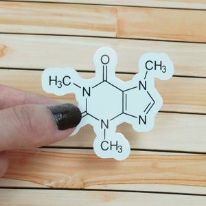 Puede incluir: Una pegatina blanca con una estructura química negra de cafeína, con el texto "H3C", "CH3", "N" y "O".