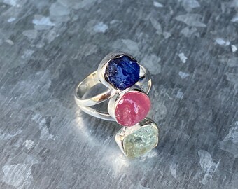 May include: A silver ring with three gemstones: a blue sapphire, a pink ruby, and a light green aquamarine.