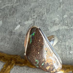 May include: A silver ring featuring a large, irregularly shaped opal. The opal has a dark brown base color with flecks of green and blue. The ring's band is a simple, polished silver.