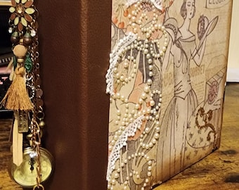 May include: A brown leather-bound journal with a vintage-inspired cover featuring a woman in a dress and a pearl necklace. The cover also includes the text "TEESDALE" and "CORSET".