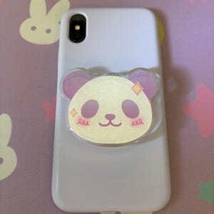 May include: A white smartphone case featuring a circular panda-shaped grip attachment. The panda face is light yellow with purple ears and eye patches, accented by small pink blush marks and two yellow diamond-shaped sparkles on its cheeks.