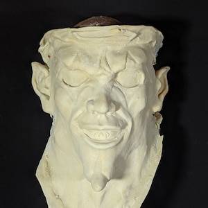 Può includere: Una maschera bianco-avorio pallido raffigurante un volto demoniaco. La maschera presenta occhi chiusi, un ampio sorriso, orecchie a punta e un mento pronunciato. La parte superiore della maschera presenta un'area strutturata e marrone, che rappresenta forse i capelli o un copricapo. La maschera è probabilmente per uso in costume o teatrale.