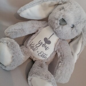 May include: A gray plush bunny with white ears and paws. The bunny has a white heart on its chest with the text "I love you Karl + Ellie" embroidered in gray.