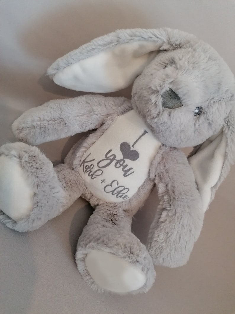 May include: A gray plush bunny with white ears and paws. The bunny has a white heart on its chest with the text "I love you Karl + Ellie" embroidered in gray.