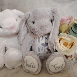 Könnte beinhalten: Zwei Plüsch-Hasen, einer grau und einer weiß, mit gestickten Namen und einem Herz auf der Brust des grauen Hasen. Der graue Hase hat den Text "Mom" und das Datum "8/8/2021" auf seiner Brust. Der weiße Hase hat den Namen "Hailee" auf einem Fuß und der graue Hase den Namen "Trey" auf dem anderen Fuß.
