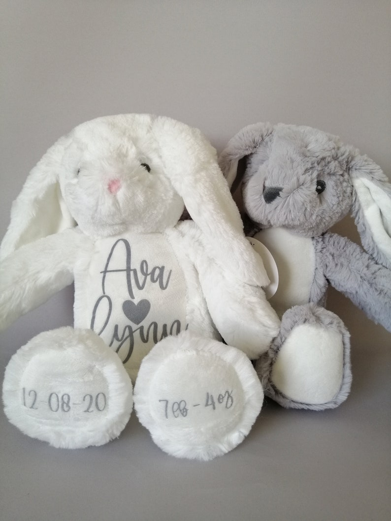 May include: Two plush bunny toys, one white and one gray, with embroidered names and dates. The white bunny has "Ava Lynn" and "12-08-20" embroidered on it, while the gray bunny has "7-08-40" embroidered on it.