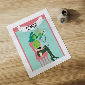 May include: A print featuring a stylized illustration of a Gemini zodiac sign. The artwork depicts a green-skinned figure seated in a pink chair, holding a hair dryer. The word "GEMINI" is displayed on a banner above the figure. The background is light blue with a pink border.