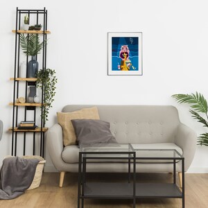 May include: A framed art print depicts a woman in a yellow dress, wearing a pink hat, and holding a drink and popcorn. The background features blue and dark blue shapes. The artwork is displayed above a gray sofa.