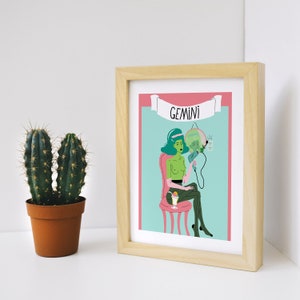 May include: A framed art print with a cartoon illustration of the Gemini zodiac sign. The artwork shows a green-skinned figure holding a mirror, seated on a pink chair, with the word "GEMINI" above. A potted cactus is to the left.