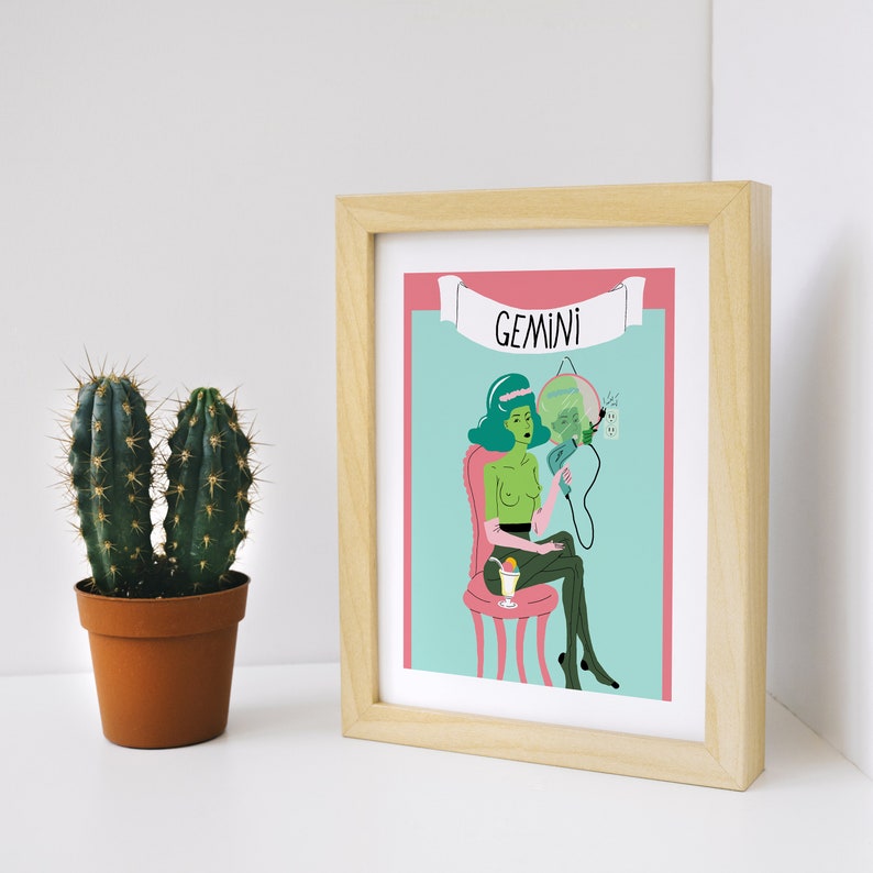 May include: A framed art print with a cartoon illustration of the Gemini zodiac sign. The artwork shows a green-skinned figure holding a mirror, seated on a pink chair, with the word "GEMINI" above. A potted cactus is to the left.