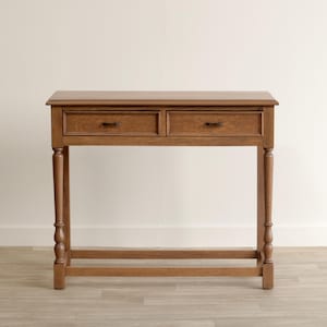 May include: A rectangular wooden console table with a medium brown finish, featuring two side-by-side drawers with small dark metal pulls. The table is supported by four slender, turned legs connected by a lower horizontal support stretcher.