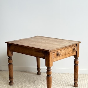 May include: An antique wooden table with a single drawer and turned legs. The table is made of light brown wood and has a natural finish. The drawer has a round wooden knob. The table is approximately 76 cm wide and 76 cm long.