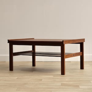 May include: A rectangular wooden coffee table with a rich brown finish, featuring a flat top surface with slightly raised edges on the sides and a lower slatted shelf supported by four sturdy legs, showcasing a clean and functional design.