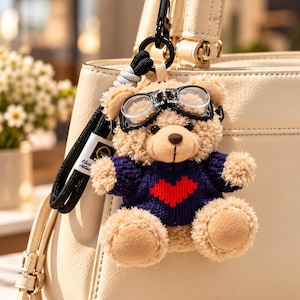 Könnte beinhalten: Ein Plüsch-Teddybär-Schlüsselanhänger mit beigem, flauschigem Körper, der einen dunkelblauen Strickpullover mit einem roten Herzmotiv auf der Vorderseite trägt. Der Bär hat eine schwarze Fliegerbrille aus Kunststoff auf dem Kopf.