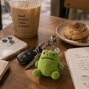 Puede incluir: Un llavero de rana verde de peluche con un cierre plateado, llaves y un teléfono sobre una mesa de madera. También se ve una taza de café con el texto "mood roastery coffee & dessert" y un bloc de notas con una lista.