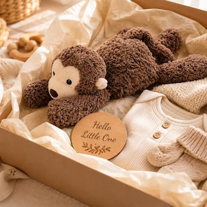 Può includere: Una scatola regalo contenente un peluche a forma di scimmia marrone, un body a maniche lunghe a coste color crema con due bottoni, un paio di scarpine in maglia e un disco in legno inciso con il testo Hello Little One e un motivo a foglie.