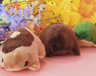 May include: Three plush toys in the shape of animals. The first is brown and tan with a brown patch on its head. The second is brown and the third is green. The toys are on a pink surface with yellow flowers in the background. The text '@practicalmagiccreations' is at the bottom of the image.