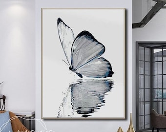 May include: A painting of a gray butterfly with blue accents, reflected in a body of water. The painting is framed in a gold frame and hangs on a white wall.