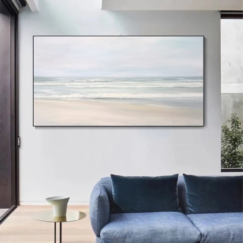May include: A framed painting of a seascape with a white sandy beach and blue ocean waves. The painting is hanging on a white wall in a living room setting.