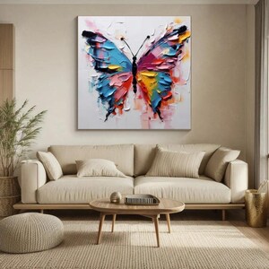 May include: A large, colorful butterfly painting with thick brushstrokes. The butterfly features blue, pink, yellow, and red hues against a white background. The artwork is displayed above a beige sofa in a living room setting.