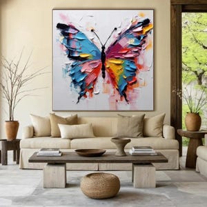 May include: Large square canvas art featuring a colorful butterfly painting. The butterfly has blue, pink, yellow, and orange wings with a black body. The artwork is displayed in a living room setting with a neutral-toned sofa and wooden furniture.