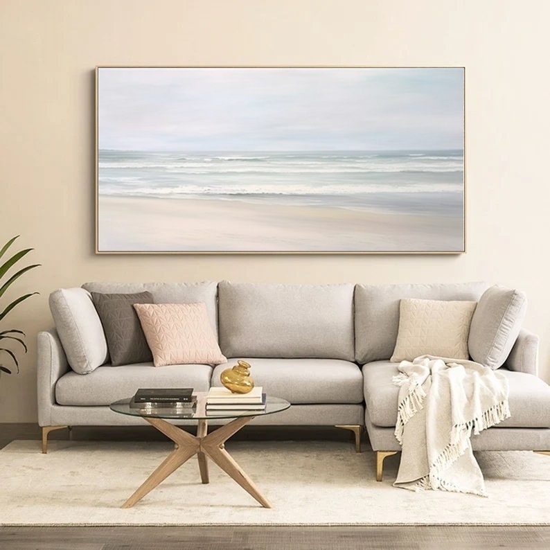 May include: A long, horizontal painting of a seascape with a white sandy beach and blue water with white waves. The painting is framed in a gold frame and is hanging on a light beige wall above a gray sectional sofa.