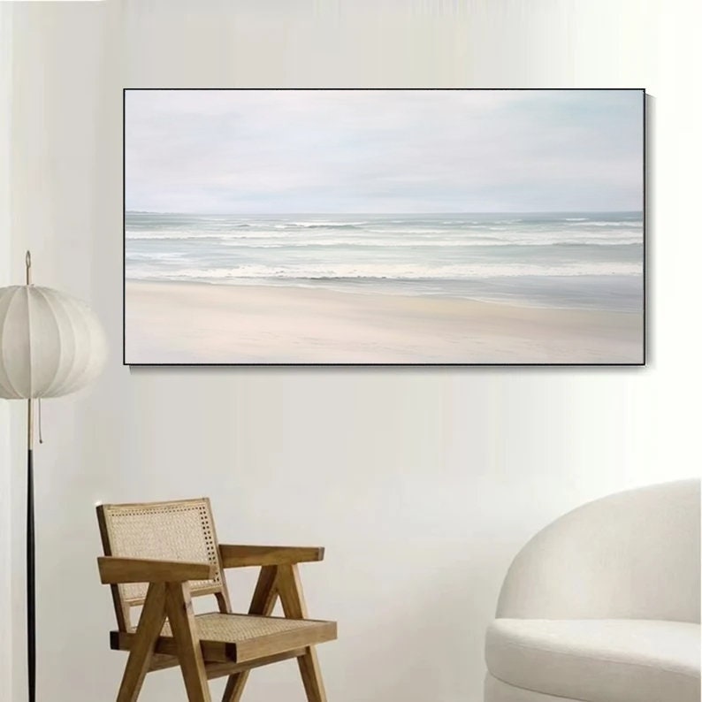 May include: A framed painting of a seascape with a white sandy beach and blue ocean waves. The painting is in a minimalist style with soft colors and a calming atmosphere.
