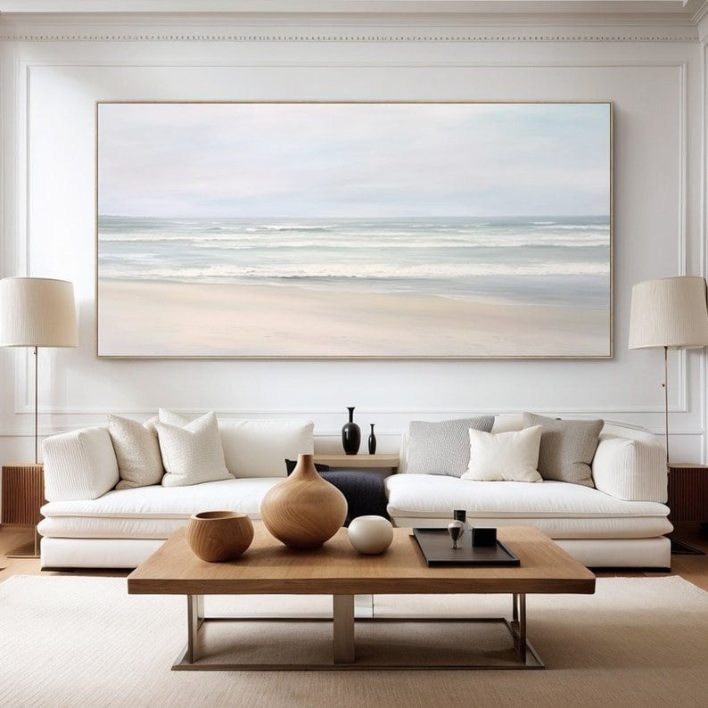 May include: A white couch with pillows sits in front of a large painting of a seascape. The painting is in shades of blue and white, with a sandy beach in the foreground. A wooden coffee table sits in front of the couch, with a wooden vase and a bowl on top.