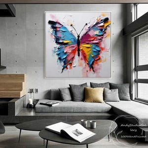 May include: A large, square, hand-painted canvas art piece featuring a colorful butterfly. The butterfly has blue, pink, yellow, and orange wings with a textured, impasto style. The artwork is set against a white background.