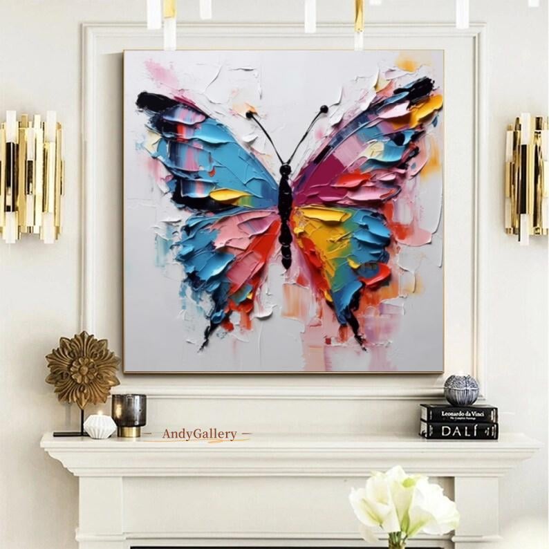 May include: A framed abstract painting of a butterfly. The butterfly's wings are rendered in thick brushstrokes of blue, pink, red, and yellow against a white background. The artwork is displayed above a white mantel.