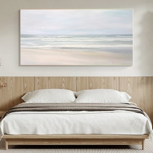 May include: A horizontal abstract painting of a beach scene with white sand and blue waves. The painting is framed in a gold frame and is hanging on a white wall above a bed.