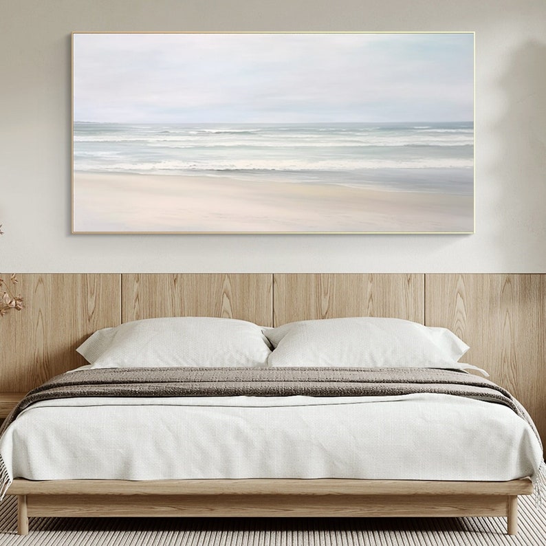 May include: A horizontal abstract painting of a beach scene with white sand and blue waves. The painting is framed in a gold frame and is hanging on a white wall above a bed.