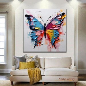 May include: A large, square canvas features a vibrant, textured painting of a butterfly. The butterfly's wings are a mix of blue, pink, yellow, and red, with a black body and antennae. The artwork is displayed above a white sofa with decorative pillows.