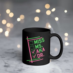 Puede incluir: Taza de cer&aacute;mica negra con las palabras "MISS MS. AKA DR." en verde y rosa, dentro de un dise&ntilde;o de lista de verificaci&oacute;n. La taza tiene un asa curva y un acabado brillante. El fondo est&aacute; borroso con luces bokeh.