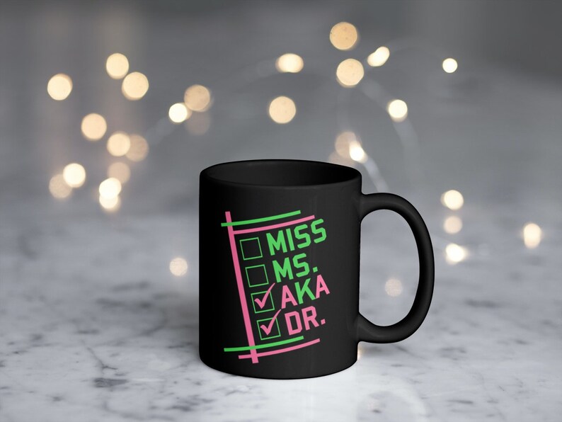 Puede incluir: Taza de cer&aacute;mica negra con las palabras "MISS MS. AKA DR." en verde y rosa, dentro de un dise&ntilde;o de lista de verificaci&oacute;n. La taza tiene un asa curva y un acabado brillante. El fondo est&aacute; borroso con luces bokeh.