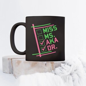 Puede incluir: Taza de cer&aacute;mica negra con las palabras "MISS MS. AKA DR." en verde y rosa. La taza tiene un asa negra y el texto est&aacute; dispuesto en formato de lista de verificaci&oacute;n. La taza est&aacute; sobre una superficie clara y texturizada.