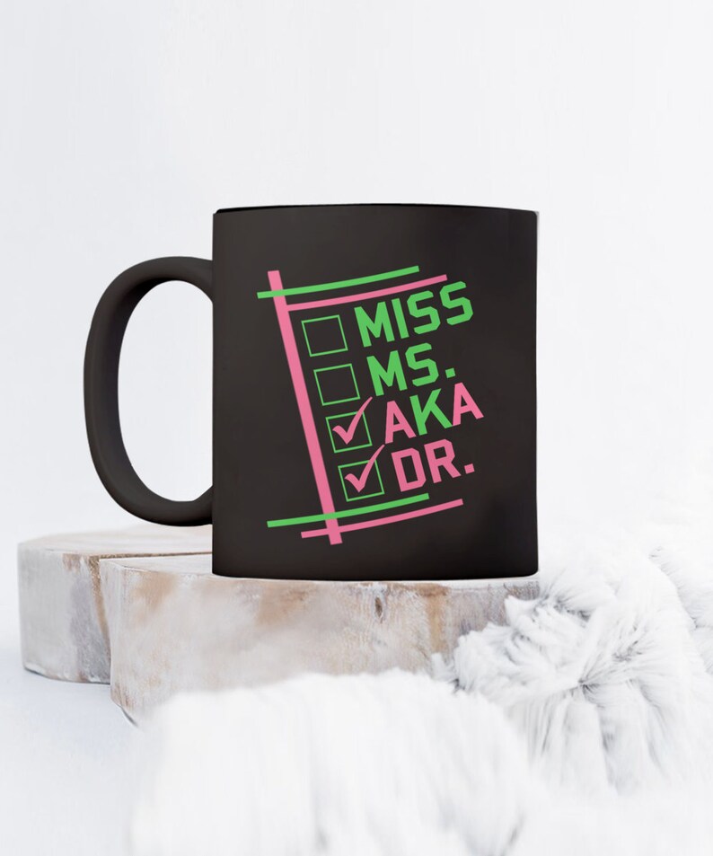 Puede incluir: Taza de cer&aacute;mica negra con las palabras "MISS MS. AKA DR." en verde y rosa. La taza tiene un asa negra y el texto est&aacute; dispuesto en formato de lista de verificaci&oacute;n. La taza est&aacute; sobre una superficie clara y texturizada.