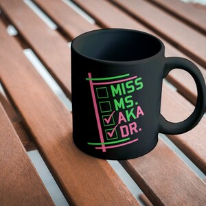 Puede incluir: Taza de cer&aacute;mica negra con las palabras "MISS MS. AKA DR." en verde y rosa. La taza tiene un asa y est&aacute; sobre una superficie de madera.