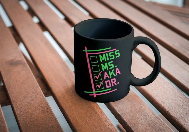Puede incluir: Taza de cer&aacute;mica negra con las palabras "MISS MS. AKA DR." en verde y rosa. La taza tiene un asa y est&aacute; sobre una superficie de madera.