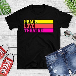 May include: Black t-shirt with the text "Peace Love Theatre" in colourful stripes. The shirt is laid out on a white wooden surface with a pair of jeans and trainers.