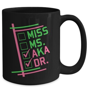 Puede incluir: Taza de cer&aacute;mica negra con las palabras "MISS MS. AKA DR." en verde y rosa. La taza presenta casillas de verificaci&oacute;n junto a "AKA" y "DR.". La taza tiene un asa curva.