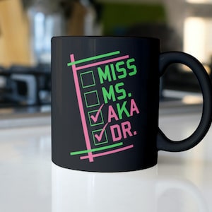Puede incluir: Taza de cer&aacute;mica negra con asa curvada. La taza presenta las palabras "MISS", "MS.", "AKA" y "DR." en verde y rosa, con casillas de verificaci&oacute;n. La taza est&aacute; sobre una superficie blanca.