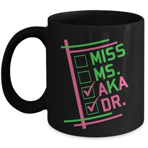 Puede incluir: Taza de cer&aacute;mica negra con asa blanca que presenta el texto "MISS MS. AKA DR." en verde y rosa. El dise&ntilde;o incluye casillas de verificaci&oacute;n y l&iacute;neas en los mismos colores. Una taza de caf&eacute; elegante y funcional.